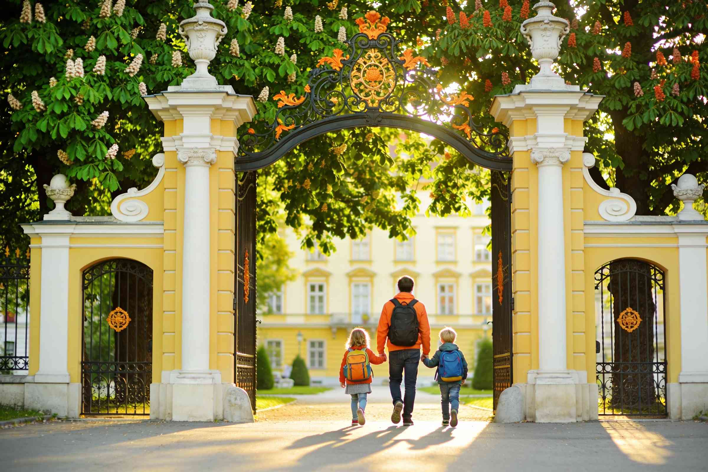 A parent and two children walking through a white baroque park gate towards the trees